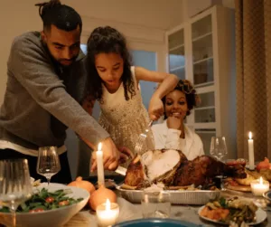 A father and daughter cut into a turkey while the family looks on. 