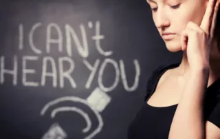 A woman holds her ear and stands before a blackboard that says, “I can’t hear you.”